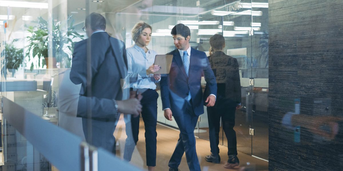 Businessman and Businesswoman Walking Through Glass Hallway, Discussing Work and Using Tablet Computer. Busy Corporate Office Building with Many Workers.